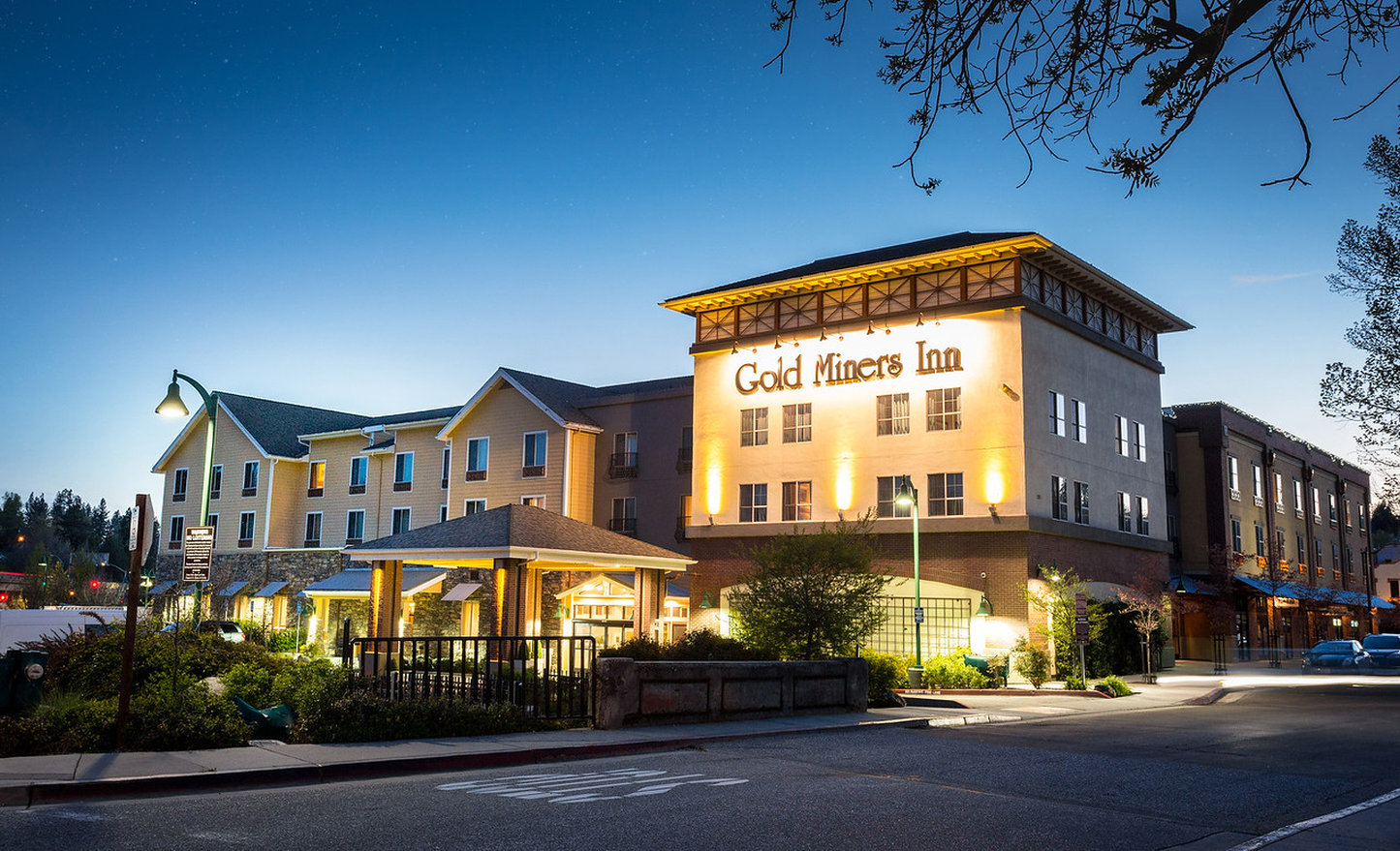 A three story hotel building appearing golden with lights shining on it at sunset. Name on building Gold Miners Inn