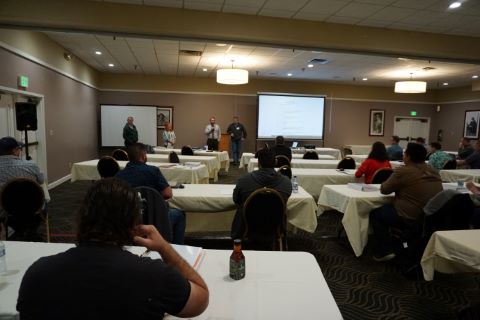 A seated audience looks to the front of a room where four people are beside a projector screen.