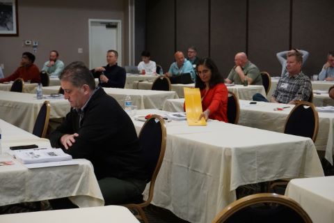 A woman holds a yellow rectangle pavement marker to see the markings, while a seated audience is listening to a speaker.