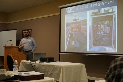Man stands at podium. Behind him is projector showing slide with image of a wrong way road sign.