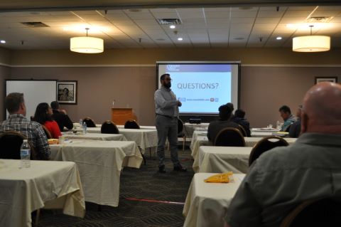 A man stands between tables with people seated as he gestures to a projector screen.