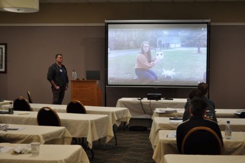 Man stands in front of projector screen. On the screen is a video call with a woman holding a drone.