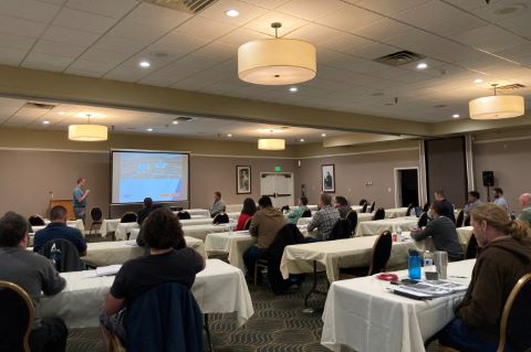 An audience seated at tables facing a projector screen and a speaker.