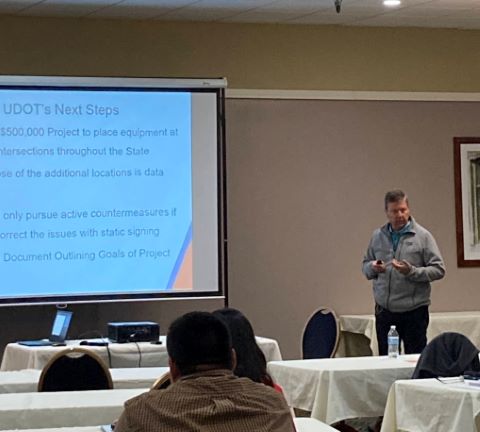 A man speaking standing to the right of a projector screen.