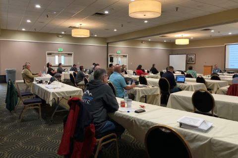 An audience seated at rectangular tables arranged classroom style. 