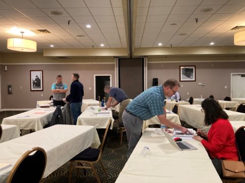Two people talk with each other while standing (left). One person points to a notebook on the table in front of a seated person.