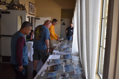 Three men stand in front of a table. They are looking at a stack of binders.