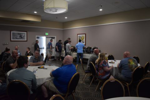 A group of people sit at tables discussing. In the background, more people stand by a buffet table.