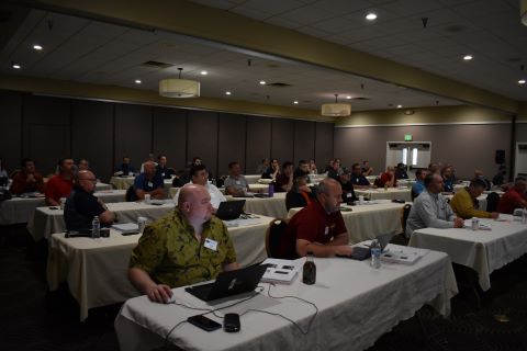 A room of people are seated at tables. They look forward at a presenter.