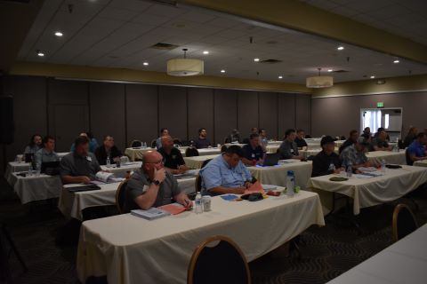 A room of people watch a presenter. In the front row, a man is taking notes.