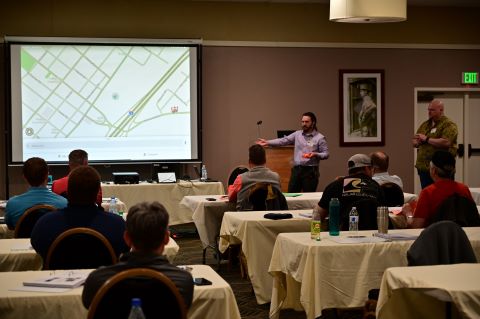 Two men stand in front of a projector. One gestures at the screen.
