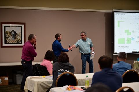Three men stand in front of an audience. The man on the right hands a microphone to the man in the middle.