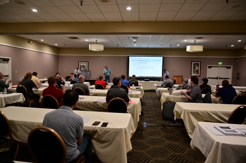 An audience watches three men standing in front of a projector screen.