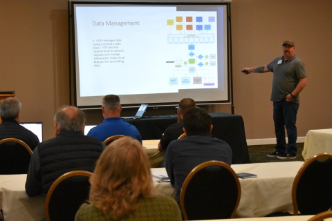 A man standing beside a projector screen points to the screen with a block diagram and text = Data Management.