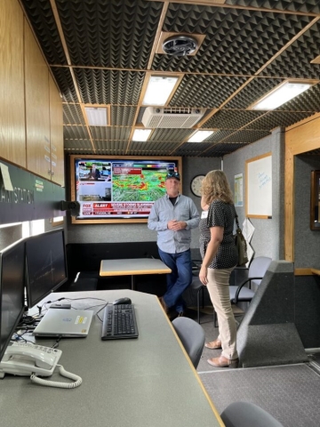 A man and a woman stand in front of a TV screen in a small room, talking to each other.