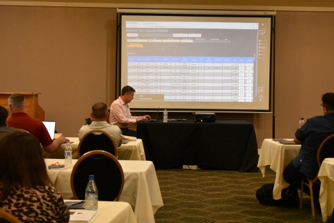 A man sits at a table in front of an audience. He is using a computer, and there is a projector screen displaying a software application data table behind him.