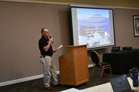 A man speaks into a microphone beside a projector screen. A mountain and some text are displayed on the screen.
