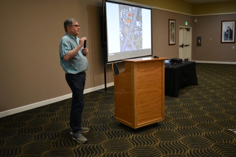 A man speaks into a microphone beside a projector screen.