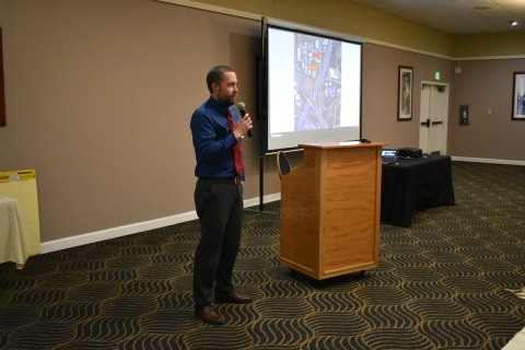 A man speaks into a microphone beside a projector screen.