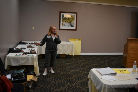 A woman speaks into a microphone and gestures at the table with speaker recognition items beside her.