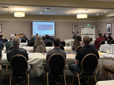 An audience seated at long tables watches a man deliver a presentation in the front of a room.
