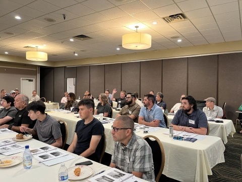 An audience seated at long tables looks towards the front of a room. A man in the center has his hand raised.