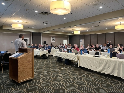A man speaks to an audience of people seated at long tables.