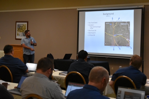 A man with a microphone gestures at a projector screen.