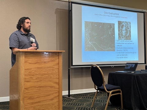 A man with a microphone stands behind a podium beside a projector screen.