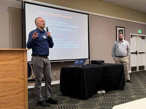 Two men stand on either side of a projector screen. The one on the left is holding a microphone and speaking to an audience.