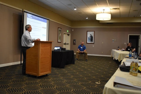 A man with a microphone presents to an audience beside a projector screen. Another man is seated across from him.