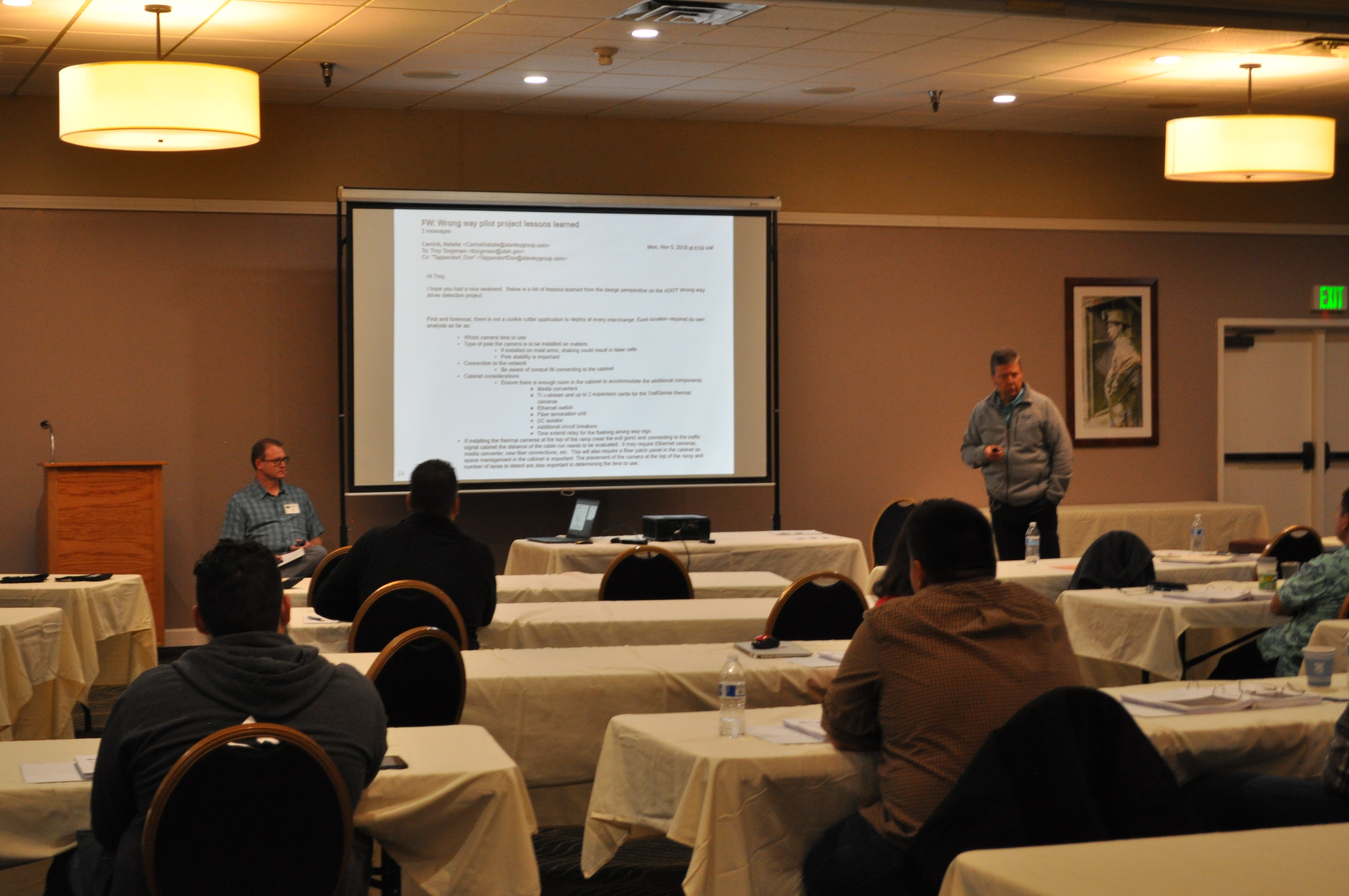 Audience members watch two men presenting on either side of a projector screen.