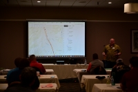 A man presents next to a projector screen with a slide showing a map.