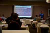 A man beside a projector screen speaks to an audience.