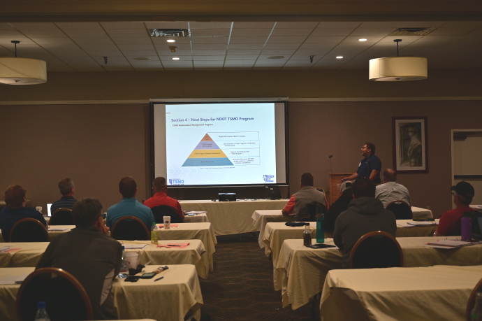 An audience watches a man present beside a projector screen with a slide showing a pyramid chart.