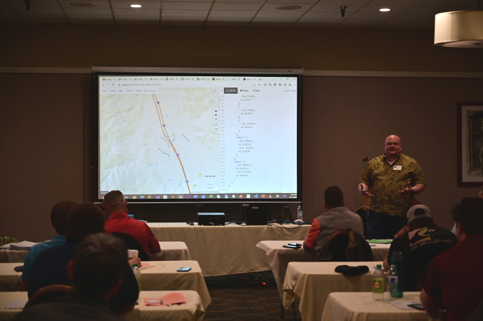 A man presents next to a projector screen with a slide showing a map.