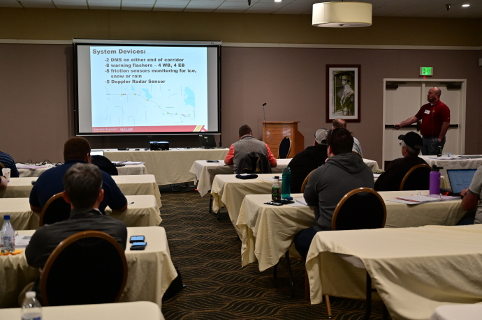 An audience watches a man presenting beside a projector screen showing a slide listing System Devices.