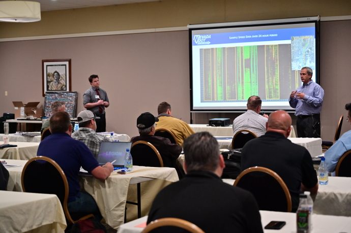 An audience watches two men presenting on either side of a projector screen showing an image of sensing results.