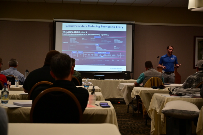A man beside a projector screen speaks to an audience.