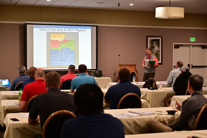 Audience members watch a man presenting beside a projector screen with a slide showing a State of Utah Map & UDOT Regions.