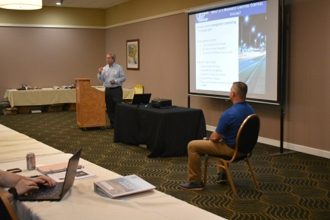 One man presents beside a projector screen. Another man is seated on the other side of the screen.