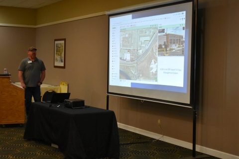 A man presents beside a projector screen showing a map and an image of a building.