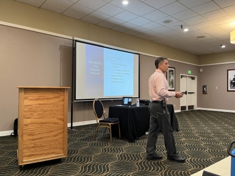 A man speaks to an audience while walking in front of a projector screen.