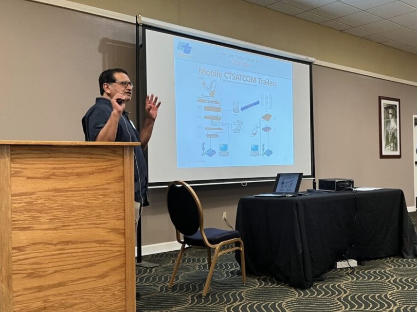 A man presents in front of a projector screen and next to a podium.