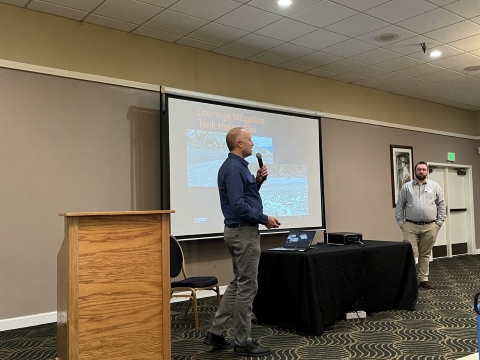 Two men stand on either side of table with a laptop and projector, screen in the background. The one on the left is holding an microphone and speaking.