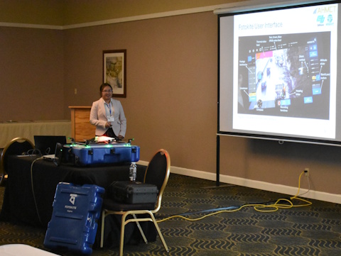 A woman stands beside a projector screen with slide showing image and 'Fotokite User Interface'. In front of her on a table are three cases and a Fotokite drone.