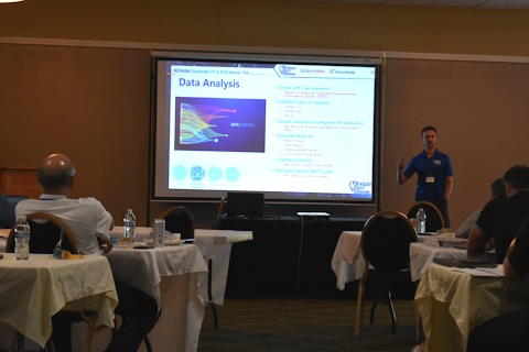 A man gestures while speaking beside a projector screen with a slide showing 'Data Analysis', graph, text; audience in front.