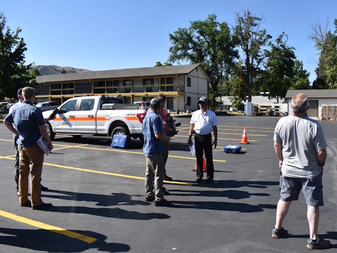In a parking lot, six men gathered in three groups talking, one on the right looks skyward, one in the center is leading discussion, DOT pickup truck and Fotokite base station in the background.