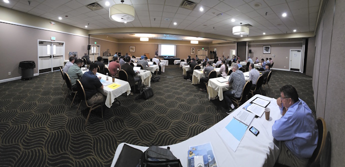 Meeting room with participants seated at tables classroom style listening to a speaker in the front of the room.
